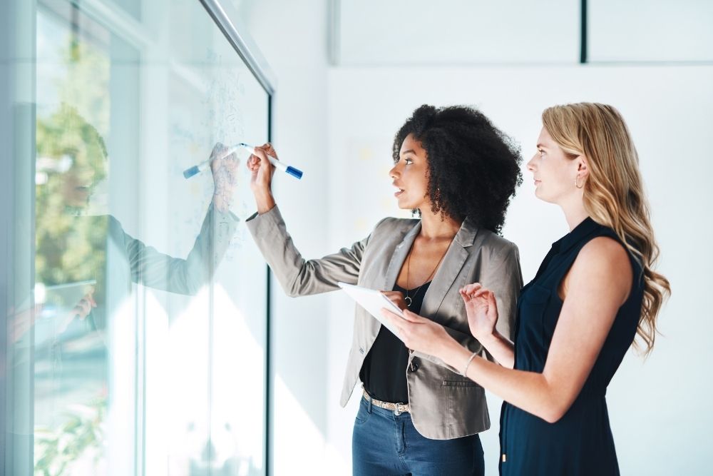 Two women discussing brand strategy on a whiteboard to adapt to the shift toward Generative Search Optimization.