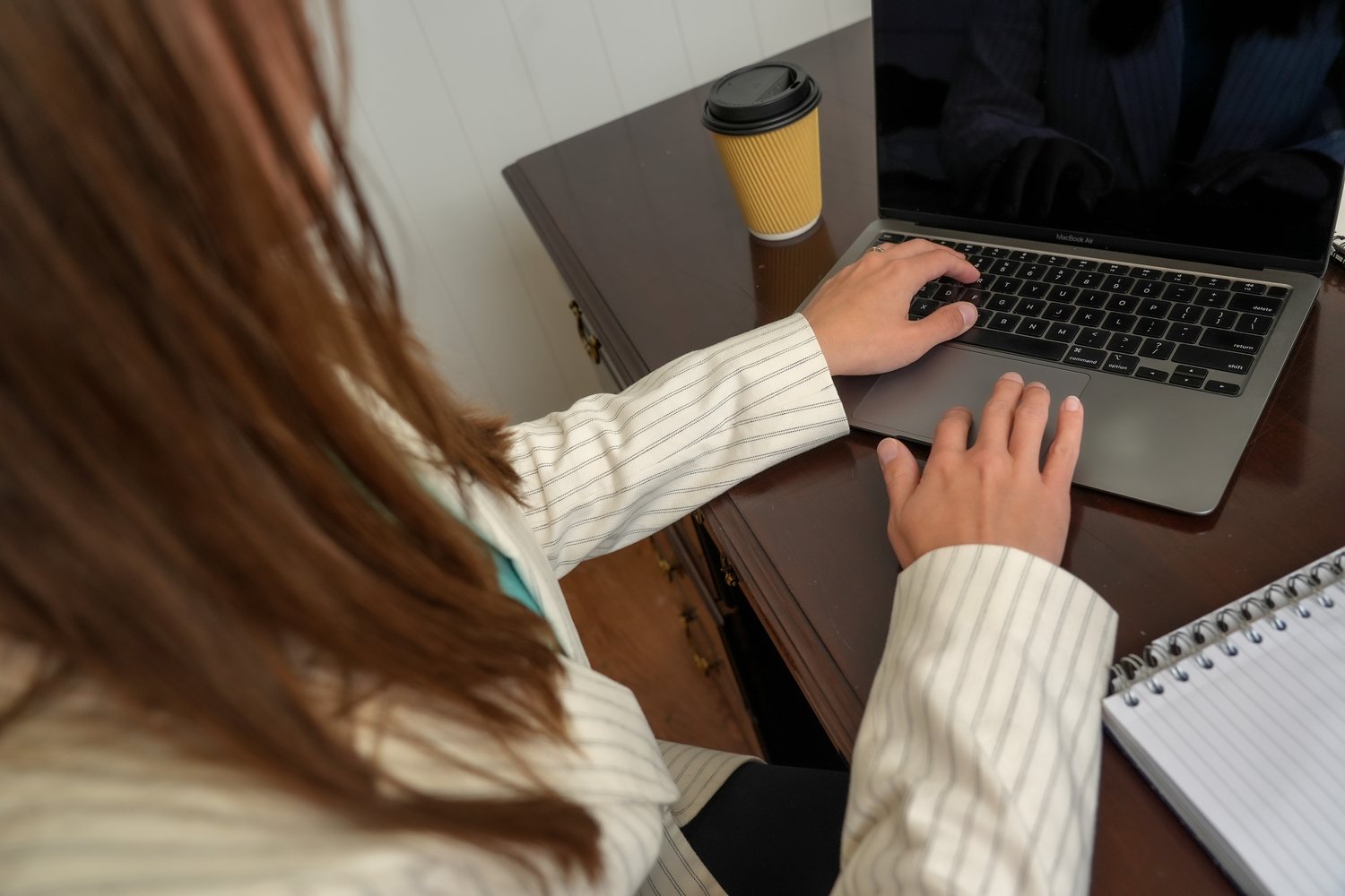 Megan typing on a laptop at the desk with a coffee and a notebook